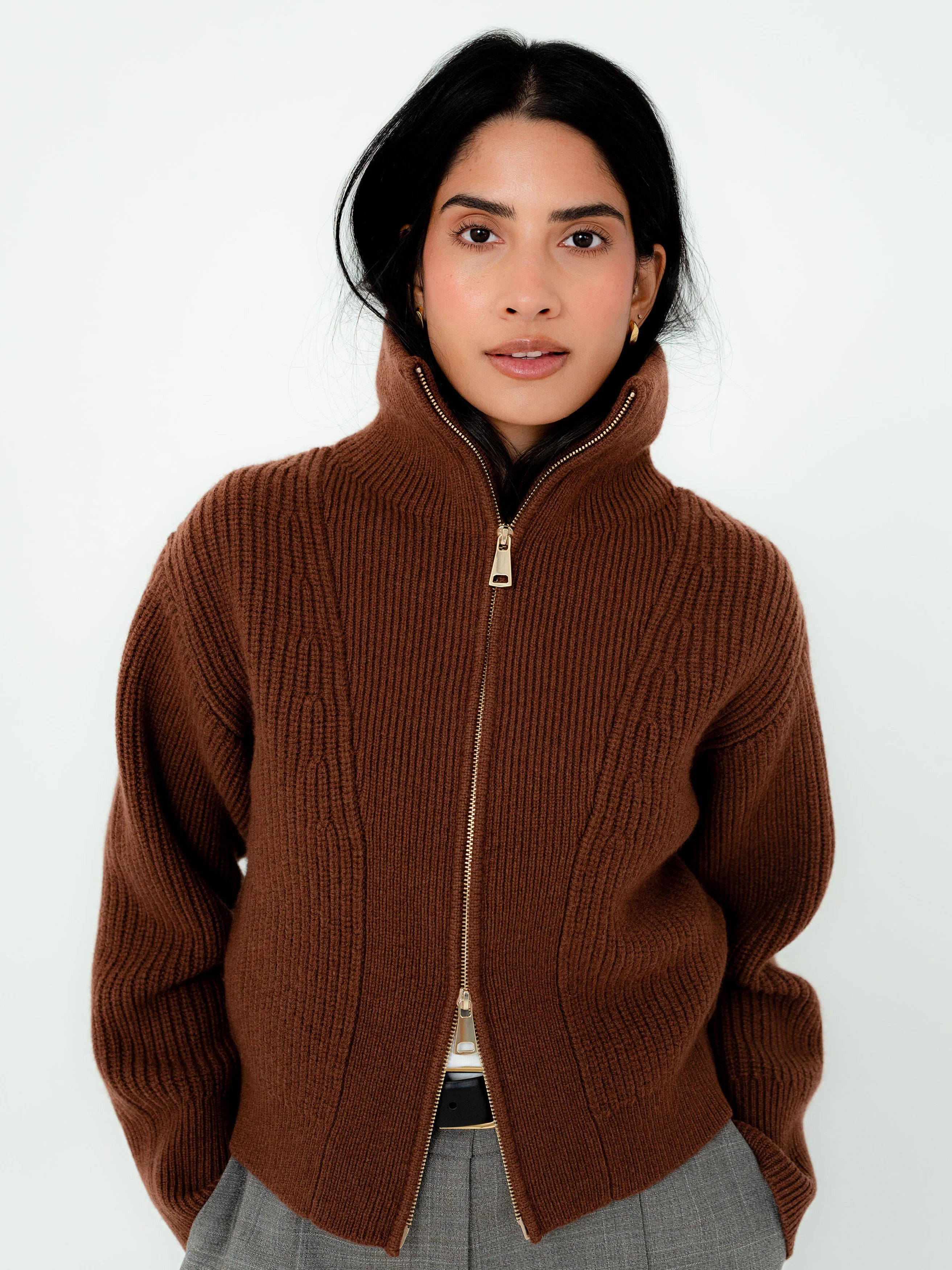 A woman wearing a brown ribbed knit zip-up sweater with a high stand-up collar, paired with gray trousers, against a plain white background.