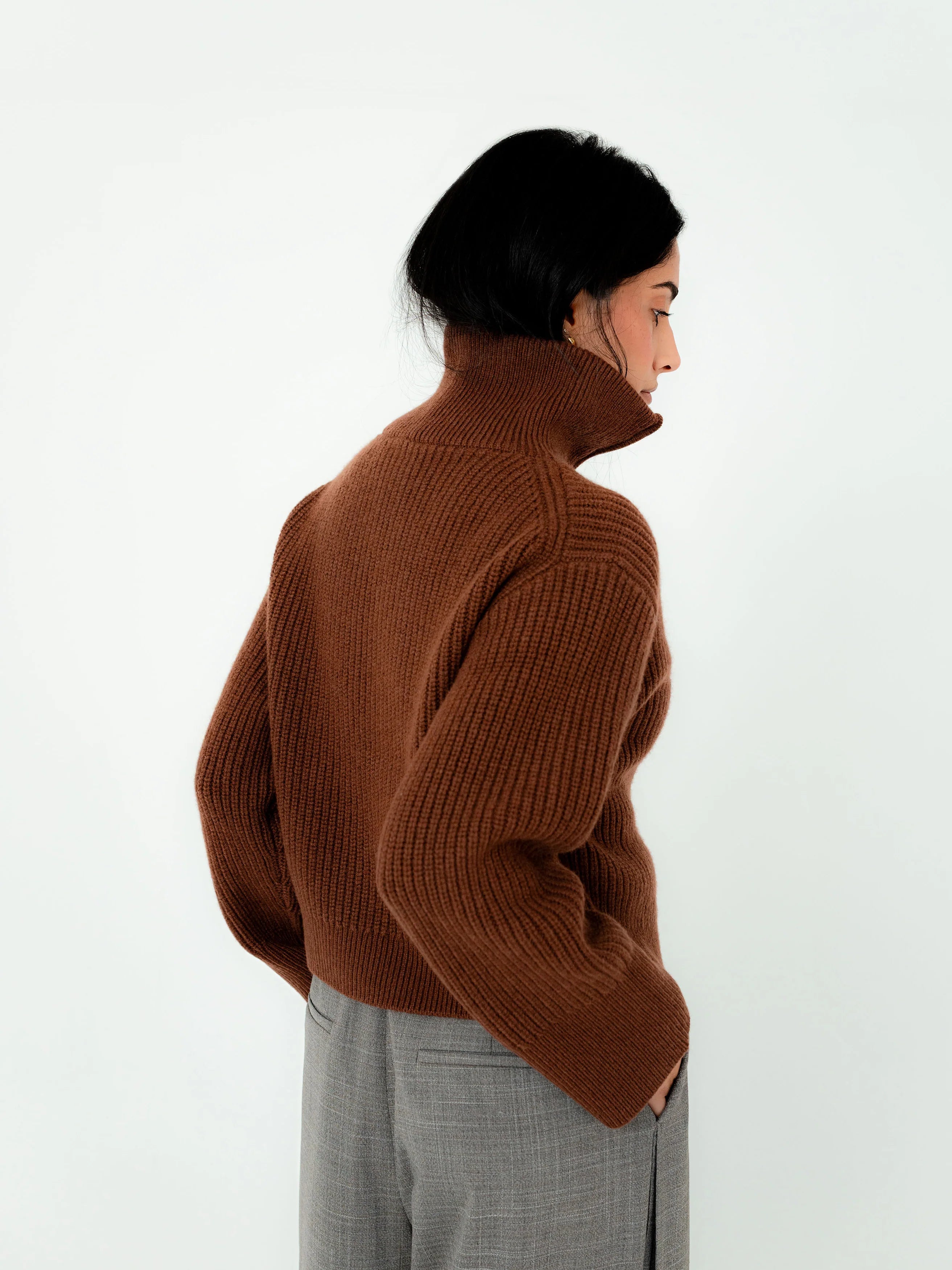 Person wearing a brown turtleneck sweater and gray pants against a white background