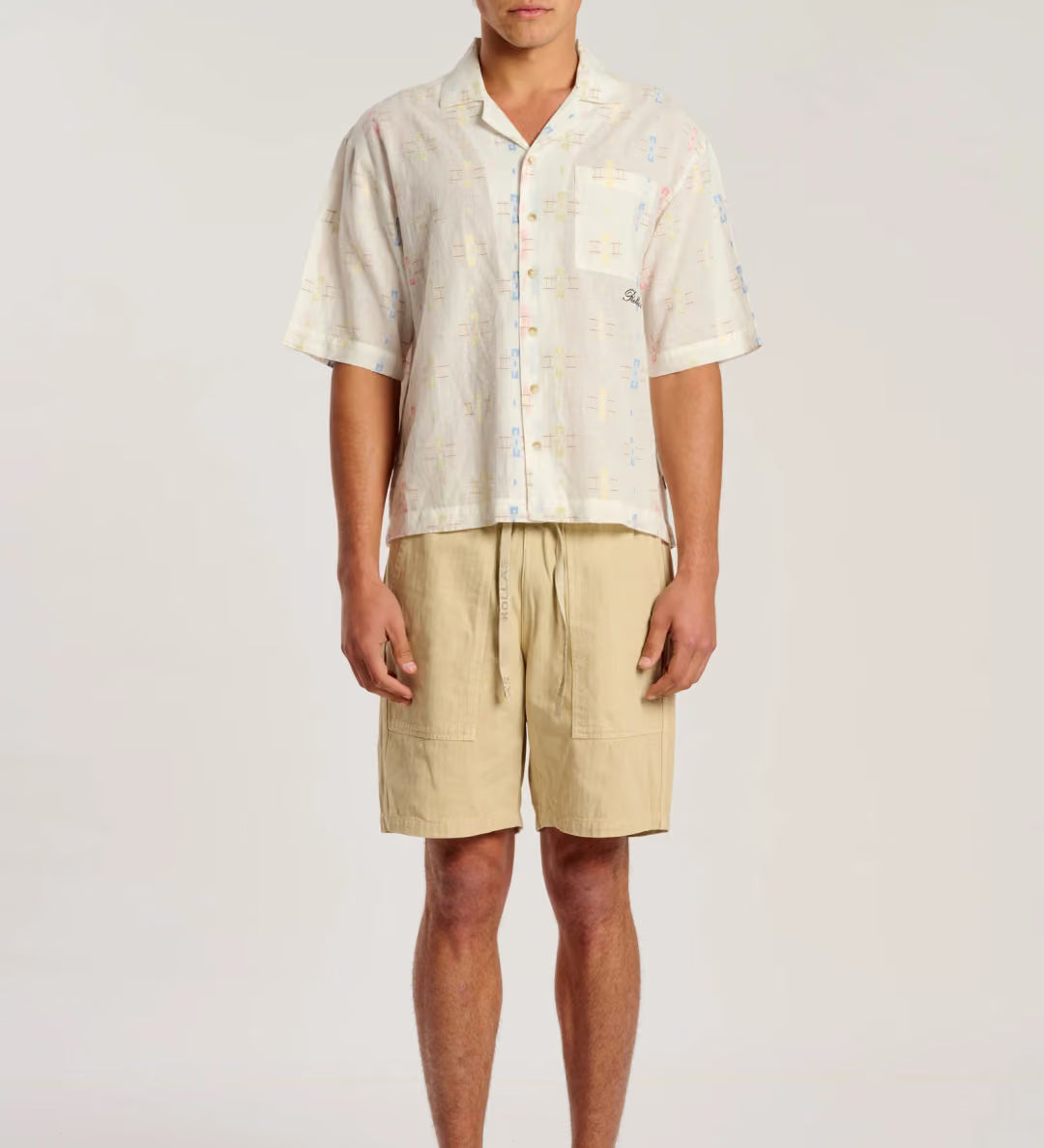 A man modeling a short-sleeve button-up shirt and beige drawstring shorts against a plain light gray studio background.