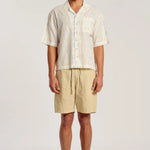 A man modeling a short-sleeve button-up shirt and beige drawstring shorts against a plain light gray studio background.