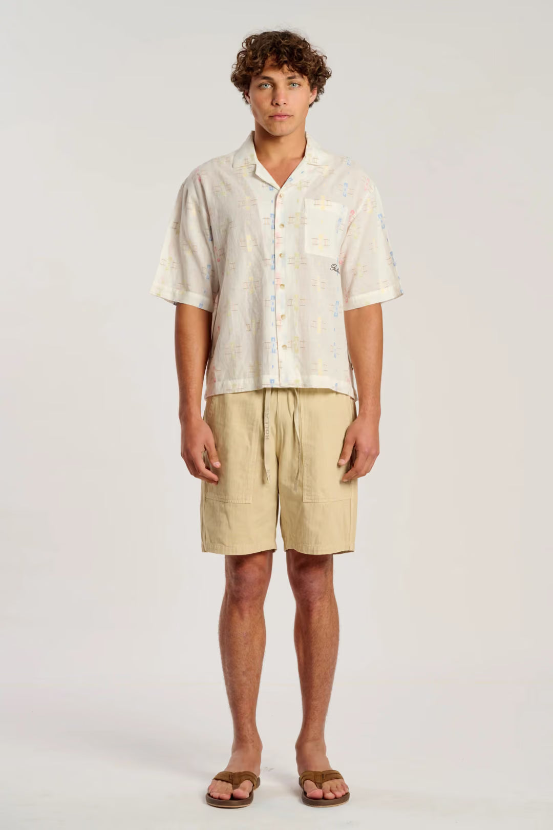 A man modeling a short-sleeve button-up shirt and beige drawstring shorts against a plain light gray studio background.