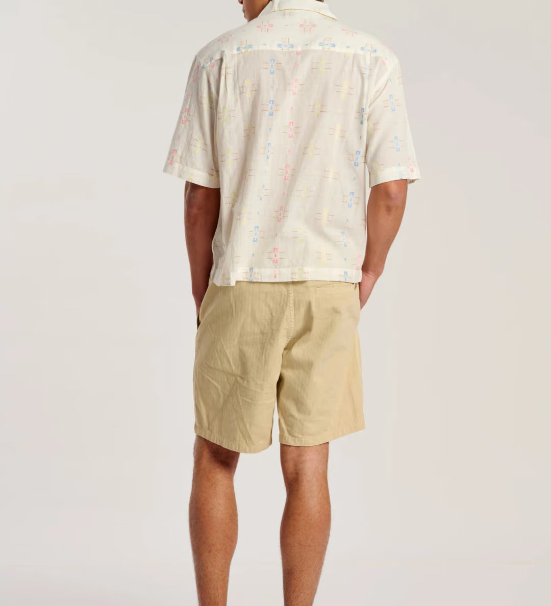 A man is shown from behind wearing a cream-colored short-sleeve button-down shirt with a subtle multicolor speckled pattern, paired with beige shorts and brown sandals, against a plain light gray studio background.