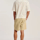 A man is shown from behind wearing a cream-colored short-sleeve button-down shirt with a subtle multicolor speckled pattern, paired with beige shorts and brown sandals, against a plain light gray studio background.