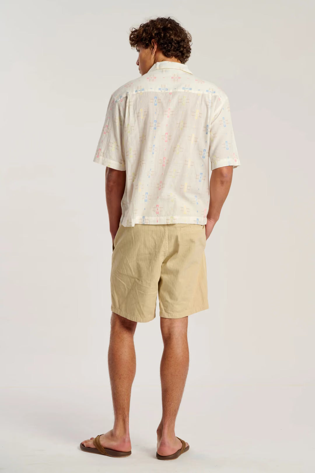 A man is shown from behind wearing a cream-colored short-sleeve button-down shirt with a subtle multicolor speckled pattern, paired with beige shorts and brown sandals, against a plain light gray studio background.