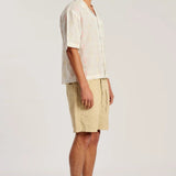 A man stands in profile wearing a light cream short-sleeve button-up shirt with a subtle pink pattern, beige shorts, and brown sandals against a plain light gray studio background.