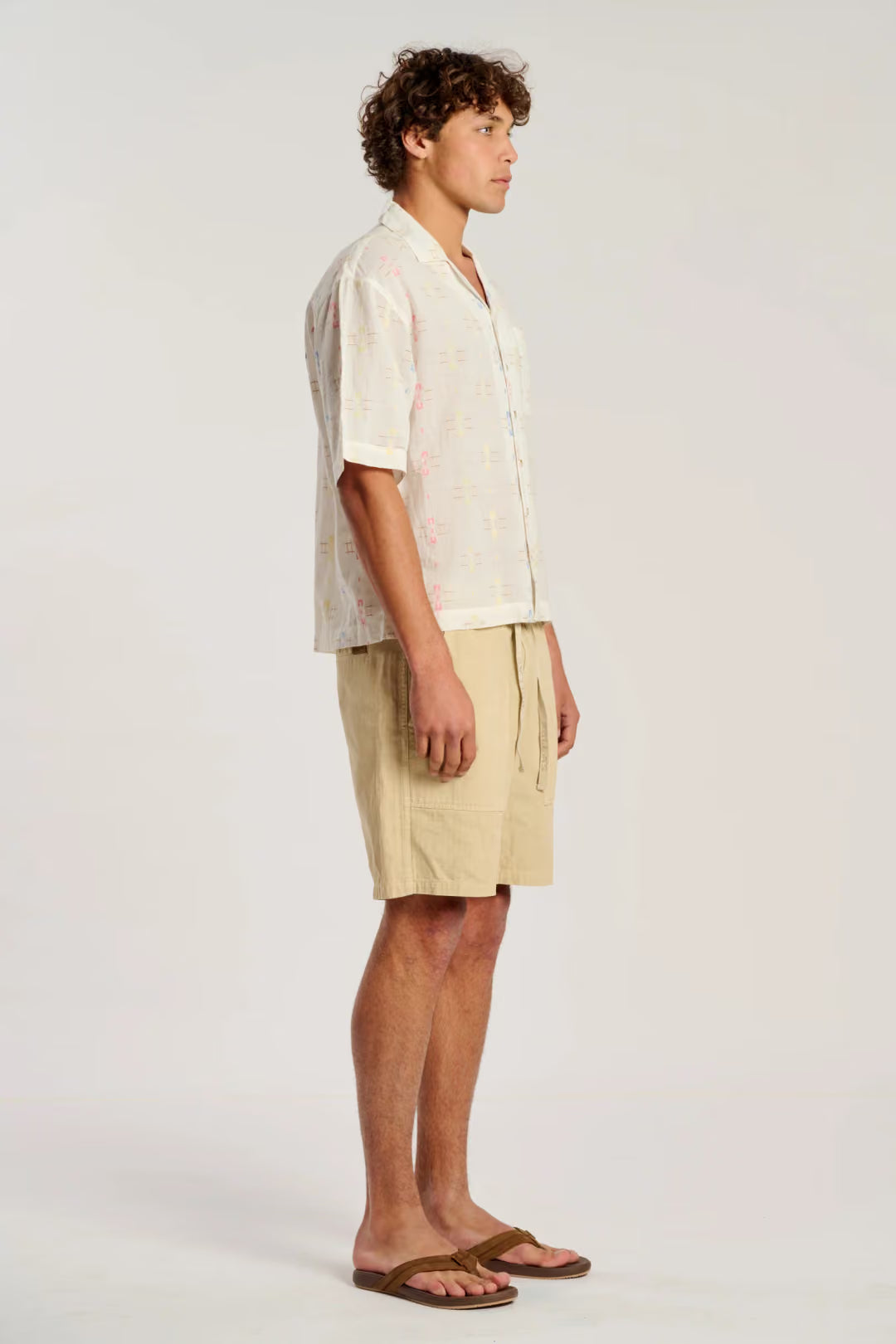 A man stands in profile wearing a light cream short-sleeve button-up shirt with a subtle pink pattern, beige shorts, and brown sandals against a plain light gray studio background.