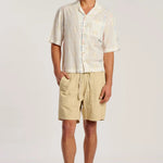 A man modeling a short-sleeve button-up shirt and beige drawstring shorts against a plain light gray studio background.