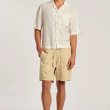 A man modeling a short-sleeve button-up shirt and beige drawstring shorts against a plain light gray studio background.