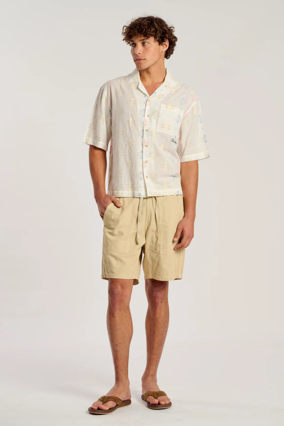 A man modeling a short-sleeve button-up shirt and beige drawstring shorts against a plain light gray studio background.