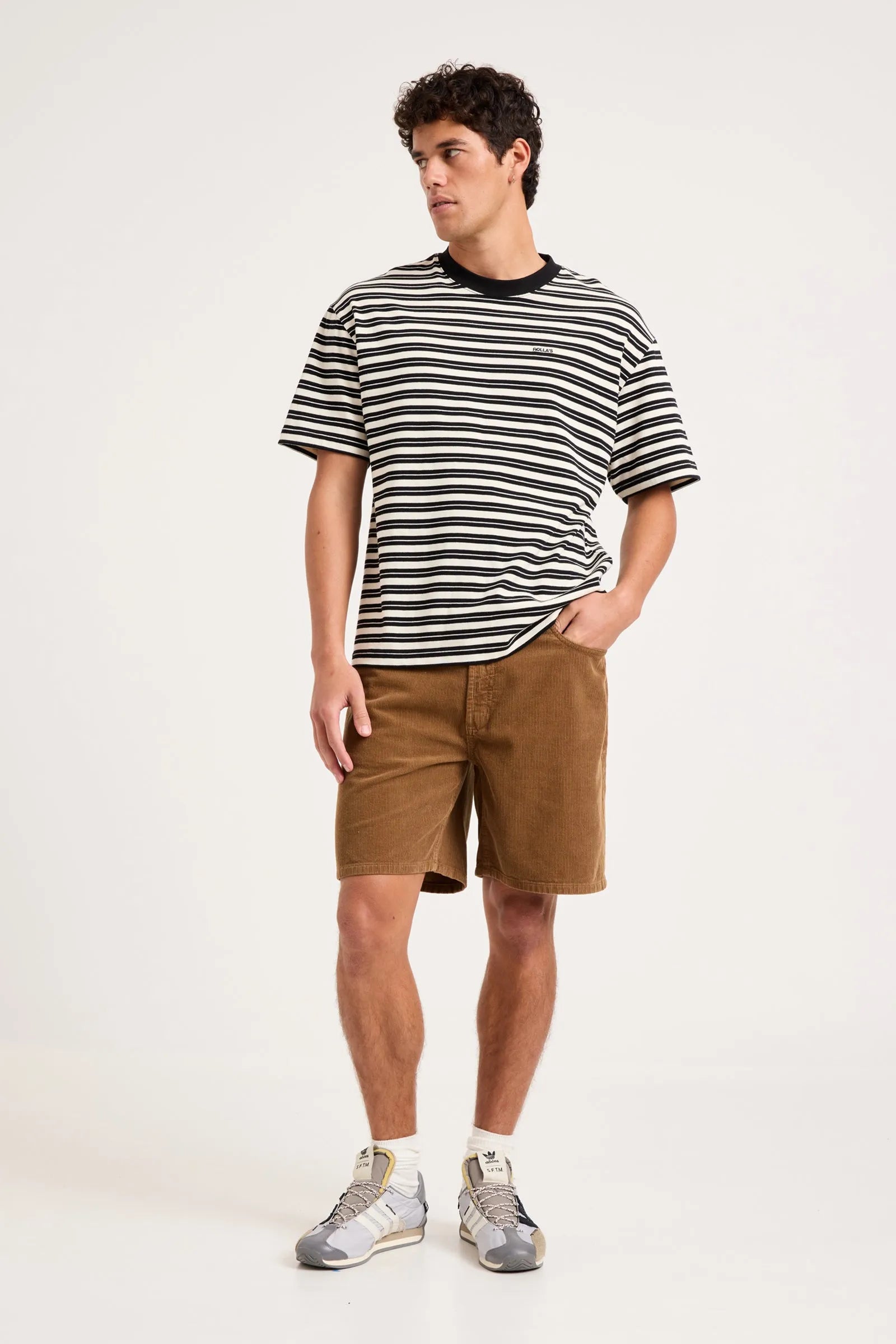 A man modeling a black and white horizontally striped short-sleeve t-shirt, brown shorts, white socks, and gray sneakers against a plain white studio background.