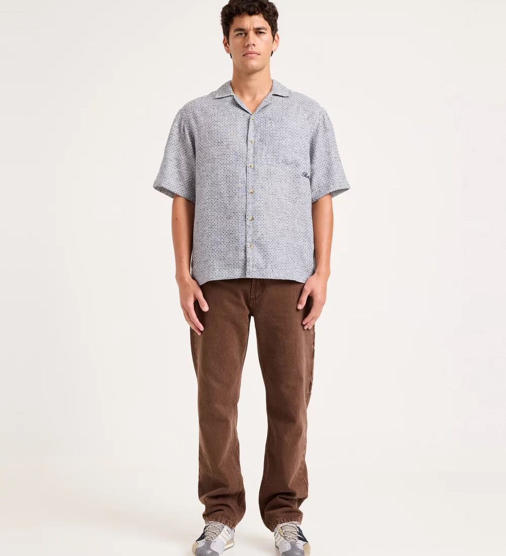 A man stands against a plain light background wearing a light gray short-sleeve button-up shirt with a subtle pattern, brown pants, and gray sneakers.