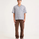 A man stands against a plain light background wearing a light gray short-sleeve button-up shirt with a subtle pattern, brown pants, and gray sneakers.