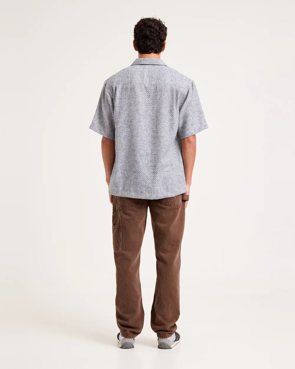A man viewed from behind wearing a light gray short-sleeve button-up shirt with a subtle textured pattern and brown pants, standing against a plain white studio background.