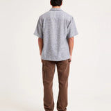 A man viewed from behind wearing a light gray short-sleeve button-up shirt with a subtle textured pattern and brown pants, standing against a plain white studio background.