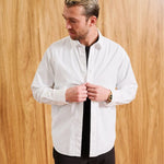 A man wearing a white long-sleeve button-down shirt with a black undershirt and black pants, standing against a light wood-paneled wall. The text 'MODEL IS 6' (183CM)' and 'SIZE MEDIUM' are visible in the bottom corners.