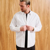 A man wearing a white long-sleeve button-down shirt with a black undershirt and black pants, standing against a light wood-paneled wall. The text 'MODEL IS 6' (183CM)' and 'SIZE MEDIUM' are visible in the bottom corners.