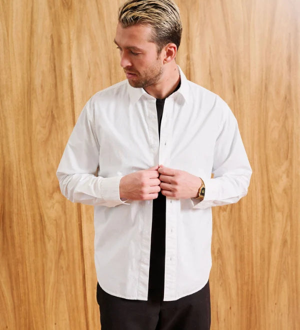 A man wearing a white long-sleeve button-down shirt with a black undershirt and black pants, standing against a light wood-paneled wall. The text 'MODEL IS 6' (183CM)' and 'SIZE MEDIUM' are visible in the bottom corners.