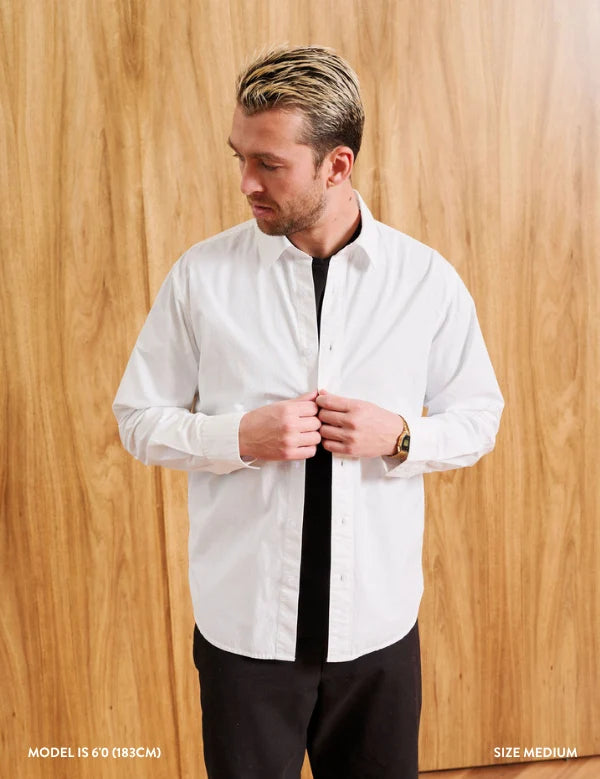 A man wearing a white long-sleeve button-down shirt with a black undershirt and black pants, standing against a light wood-paneled wall. The text 'MODEL IS 6' (183CM)' and 'SIZE MEDIUM' are visible in the bottom corners.