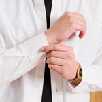 A close-up of a man wearing a white long-sleeve button-down shirt over a black undershirt, adjusting the cuff of the shirt. He is wearing a gold watch on his wrist.