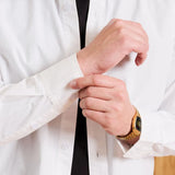 A close-up of a man wearing a white long-sleeve button-down shirt over a black undershirt, adjusting the cuff of the shirt. He is wearing a gold watch on his wrist.