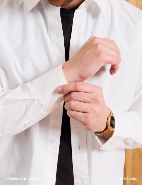A close-up of a man wearing a white long-sleeve button-down shirt over a black undershirt, adjusting the cuff of the shirt. He is wearing a gold watch on his wrist.