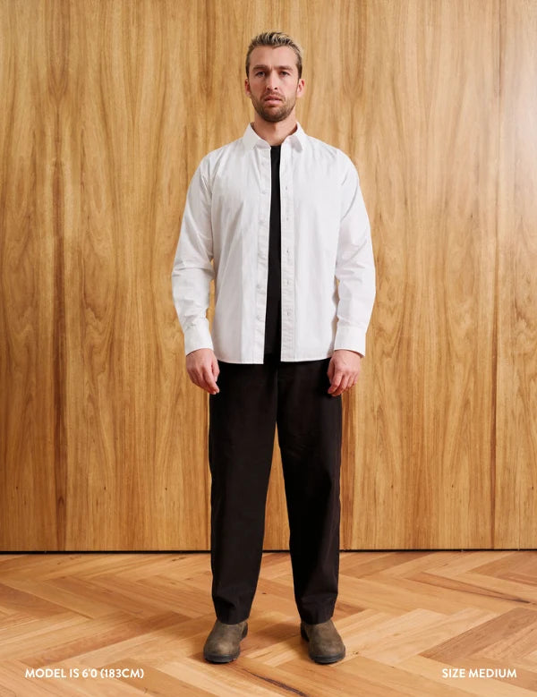 A man stands against a wood-paneled wall wearing a white long-sleeve button-up shirt, a black tie, and black trousers. Text at the bottom reads 'MODEL 18-60 (183CM)' and 'SIZE MEDIUM'.