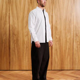 A man modeling a white long-sleeve button-down shirt with a black tie and black trousers, standing against a light wood-paneled wall and wood floor. Text overlays at the bottom indicate 'MODEL 15' (6'0" / 183CM) and 'SIZE MEDIUM'.