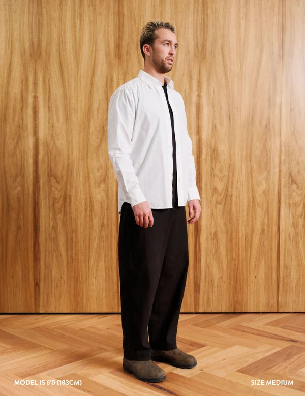 A man modeling a white long-sleeve button-down shirt with a black tie and black trousers, standing against a light wood-paneled wall and wood floor. Text overlays at the bottom indicate 'MODEL 15' (6'0" / 183CM) and 'SIZE MEDIUM'.