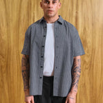 A man wearing a short-sleeve black and white gingham button-up shirt over a white t-shirt and black pants, standing against a wood-paneled wall. Text at the bottom reads 'MODEL IS 5'9 (177CM)' and 'SIZE MEDIUM'.