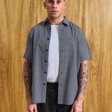 A man wearing a short-sleeve black and white gingham button-up shirt over a white t-shirt and black pants, standing against a wood-paneled wall. Text at the bottom reads 'MODEL IS 5'9 (177CM)' and 'SIZE MEDIUM'.