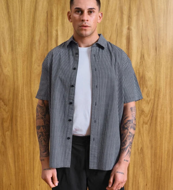 A man wearing a short-sleeve black and white gingham button-up shirt over a white t-shirt and black pants, standing against a wood-paneled wall. Text at the bottom reads 'MODEL IS 5'9 (177CM)' and 'SIZE MEDIUM'.