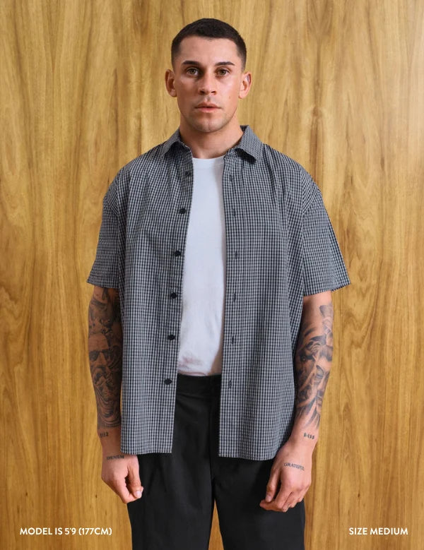 A man wearing a short-sleeve black and white gingham button-up shirt over a white t-shirt and black pants, standing against a wood-paneled wall. Text at the bottom reads 'MODEL IS 5'9 (177CM)' and 'SIZE MEDIUM'.