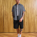 A man modeling a short-sleeve button-up shirt with a fine gray and white checkered pattern, worn over a white t-shirt and paired with black shorts, white socks, and black loafers, standing against a wood-paneled wall.
