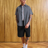 A man modeling a short-sleeve button-up shirt with a fine gray and white checkered pattern, worn over a white t-shirt and paired with black shorts, white socks, and black loafers, standing against a wood-paneled wall.