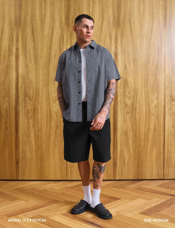 A man modeling a short-sleeve button-up shirt with a fine gray and white checkered pattern, worn over a white t-shirt and paired with black shorts, white socks, and black loafers, standing against a wood-paneled wall.