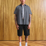 A man stands in a room with a wood-paneled wall and wood floor, wearing a short-sleeve black and white gingham button-up shirt over a white undershirt, black shorts, white socks, and black loafers. Text at the bottom reads 'MODEL 18'9' (177CM)' and 'SIZE MEDIUM'.