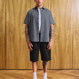 A man stands in a room with a wood-paneled wall and wood floor, wearing a short-sleeve black and white gingham button-up shirt over a white undershirt, black shorts, white socks, and black loafers. Text at the bottom reads 'MODEL 18'9' (177CM)' and 'SIZE MEDIUM'.