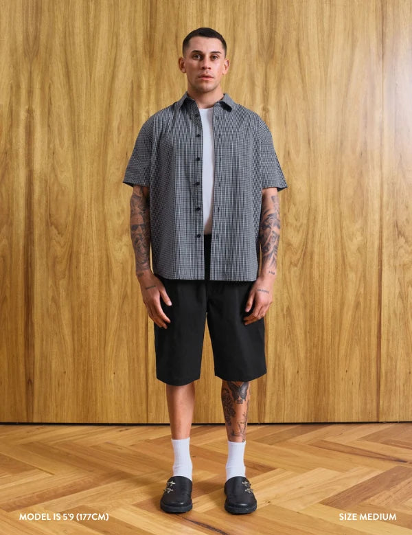 A man stands in a room with a wood-paneled wall and wood floor, wearing a short-sleeve black and white gingham button-up shirt over a white undershirt, black shorts, white socks, and black loafers. Text at the bottom reads 'MODEL 18'9' (177CM)' and 'SIZE MEDIUM'.
