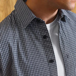 Close-up of a man wearing a black and white gingham check button-down shirt over a white t-shirt, with a light wood background.