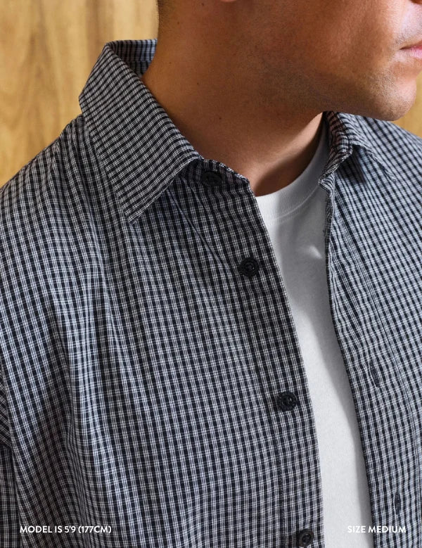 Close-up of a man wearing a black and white gingham check button-down shirt over a white t-shirt, with a light wood background.