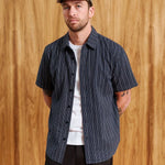 A man wearing a navy blue short-sleeve button-up shirt with thin white vertical pinstripes, layered over a white t-shirt and black pants, standing against a wooden wall background.