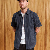 A man wearing a navy blue short-sleeve button-up shirt with thin white vertical pinstripes, layered over a white t-shirt and black pants, standing against a wooden wall background.