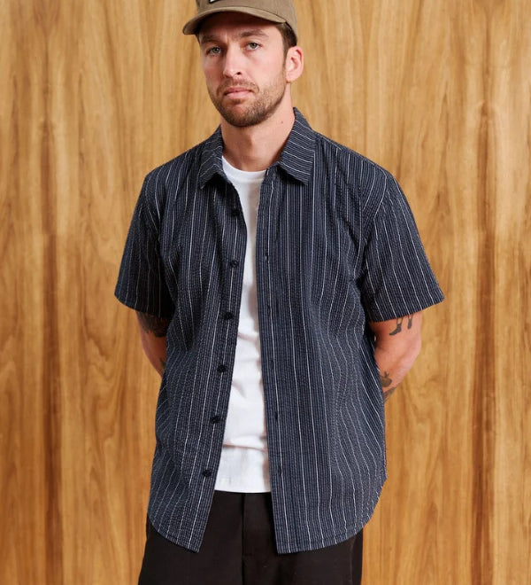 A man wearing a navy blue short-sleeve button-up shirt with thin white vertical pinstripes, layered over a white t-shirt and black pants, standing against a wooden wall background.