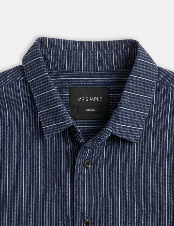 Close-up of a navy blue button-down shirt with thin white vertical pinstripes, featuring a black neck label that reads 'MR SIMPLE'.