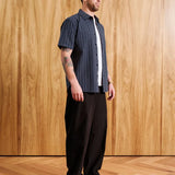 A man wearing a navy blue short-sleeve button-up shirt with thin white vertical pinstripes, black trousers, and a brown cap, standing in front of a light wood-paneled wall.