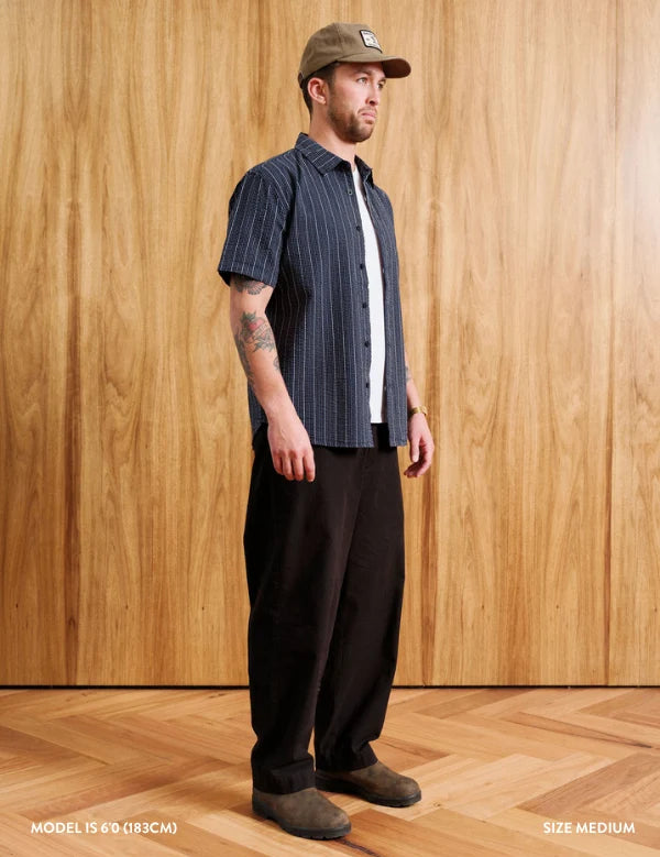 A man wearing a navy blue short-sleeve button-up shirt with thin white vertical pinstripes, black trousers, and a brown cap, standing in front of a light wood-paneled wall.