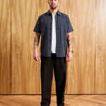 A man stands against a wood-paneled wall wearing a dark navy short-sleeve button-up shirt with thin white vertical stripes, a white t-shirt underneath, dark brown pants, and a brown cap. Text at the bottom indicates model height and size medium.