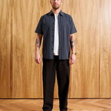 A man stands against a wood-paneled wall wearing a dark navy short-sleeve button-up shirt with thin white vertical stripes, a white t-shirt underneath, dark brown pants, and a brown cap. Text at the bottom indicates model height and size medium.