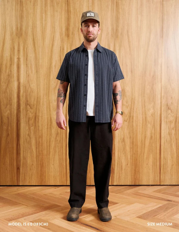 A man stands against a wood-paneled wall wearing a dark navy short-sleeve button-up shirt with thin white vertical stripes, a white t-shirt underneath, dark brown pants, and a brown cap. Text at the bottom indicates model height and size medium.