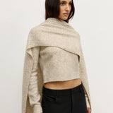 Woman wearing a beige knitted shawl over a cropped top and black pants on a white background